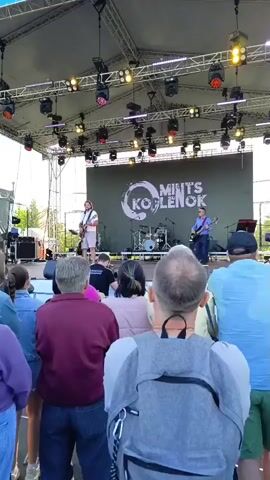 This is a wide shot of an outdoor concert. A band of at least three members is performing on a stage. Two guitarists are visible, one on the left and one on the right, with a drummer behind them. The band's name, "MINTS KOBLENOK," is displayed on a large screen behind them. The stage is equipped with numerous lights, and the area in front of the stage is filled with an audience watching the performance. The weather appears to be fair.
