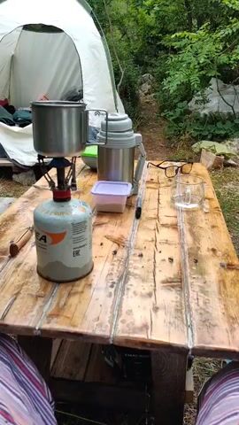 The video shows a close-up view of a portable camping stove heating a pot outdoors. The stove is connected to a propane canister and the blue flame is visible. The pot is made of metal and appears to be used for cooking or boiling water.

In the background, a white tent is partially visible, suggesting a camping or outdoor setting. The scene is set on a rustic wooden table, with some green foliage in the background, indicating a natural environment. There's also a thermos, a small plastic container, a pair of glasses, and a glass mug on the table. The lighting suggests it might be daytime, possibly morning or afternoon, with natural light filtering through the trees. The overall mood is one of outdoor leisure or preparation for a meal in nature.

Visible text on the propane canister includes "Gas Camping", "PROPANE", and "EAC". The tent has the brand "DOD" visible on it. The table has "KAKAO-BOBOK" written on it. The location context provided is Utios, Ukraine.