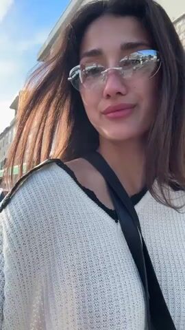 A woman with long brown hair and wearing frameless sunglasses, a white sweater with a black trim, and a black cross-body strap walks down a street. The street has white buildings on the side, and a car is parked nearby. A man's voice asks a question in the background. The woman smiles and looks at the camera. The sky is blue and sunny.
