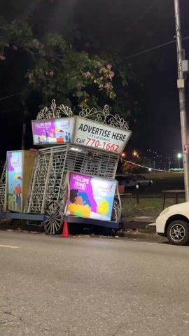 The video shows a stationary advertisement structure placed on the side of a road. The structure appears to be designed to resemble a decorative carriage or cart, with ornate metalwork and wooden wheels. It has multiple advertising panels displaying the words "WELCOME HOME!" and other colorful images. One of the larger panels reads "ADVERTISE HERE" and lists a phone number: "770-1662." A small orange traffic cone sits at the base of the structure. The background is dark, suggesting nighttime. There are streetlights visible, and a bridge-like structure with lights can be seen in the distance. A white car is parked beside the advertisement structure. A tree with some pink blossoms is partially visible in the upper left corner of the frame.