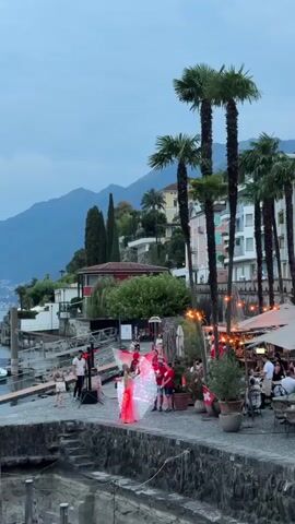 This is a vertical video captured at dusk or early evening on a waterfront promenade. In the foreground, there's a stone embankment leading down to water, with more stone steps visible. The promenade is paved and lined with potted plants and outdoor seating for a restaurant, indicated by umbrellas and tables with people. To the left, a section of the promenade appears to be a performance area, featuring a DJ setup with speakers and lighting. In the center, two individuals are performing; one is wearing a bright pink, illuminated costume with what appear to be large, flowing wings, while another person, dressed in a red shirt, is standing nearby. Several other people are scattered around the performance area and the restaurant seating. Palm trees are prominently featured along the promenade, and in the background, there are buildings nestled on a hillside overlooking a body of water, with mountains forming the backdrop under a cloudy sky. Swiss flags are visible on some of the tables.