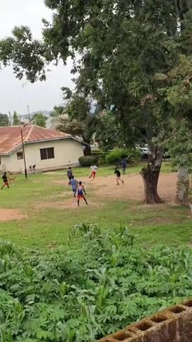 A group of children are playing soccer on a grassy field. The field is dirt and grass, with some trees and bushes around it. In the background, there is a building with a rusty roof. The sky is overcast, suggesting it might be a cloudy day. The children are wearing casual clothes and appear to be enjoying themselves. One child in the foreground is wearing a blue shirt and red shorts, while another is in a white shirt and red shorts. They are all actively engaged in the game, running and kicking the ball. The overall scene depicts a casual outdoor activity in what appears to be a residential or school setting in Anguldi, Nigeria. There are no visible text elements in the image.