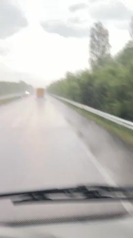 The video shows a view from inside a vehicle driving on a wet highway during a rainstorm. Raindrops are visible on the windshield. In front, a yellow truck is driving in the distance. To the left, a green truck is passing the vehicle. The road is wet and reflects the overcast sky. Trees line the sides of the highway.