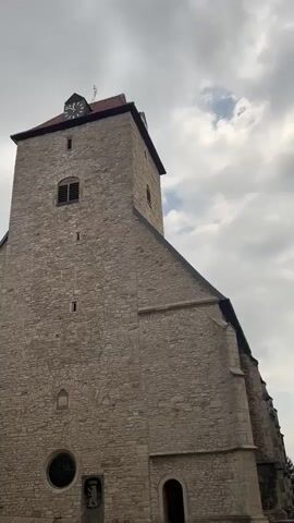 This media file is a low-angle shot of a stone church tower against a cloudy sky. The tower is tall and imposing, constructed from rough-hewn stones. It features a red-tiled roof and a clock face on one side, with Roman numerals and black hands. Below the clock, there is a narrow, arched window. Further down the tower, there are several smaller rectangular windows. At the base of the tower, there is a circular stained-glass window above a small stone carving depicting a heraldic crest with a lion. To the right of the main tower, the side of the church is visible, also made of stone, with a sloping roofline. A dark, arched doorway is visible at the bottom right of the frame. The sky is filled with dramatic, grey clouds, suggesting overcast or potentially stormy weather. The lighting is diffused, indicating it is daytime, possibly late afternoon or early morning given the cloud cover. The overall mood is somewhat somber due to the weather and the stoic nature of the stone architecture. No people are visible in the frame. The location is identified as Schöningen, Germany. There is no visible text in the media file itself.