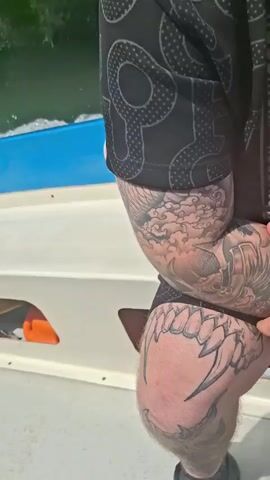 A boat speeds through a waterway lined with lush green trees. The boat is white with a blue trim and several red and white circular stickers on its side. The camera is positioned on the boat, partially focusing on a man with numerous tattoos on his arm and legs. He's wearing a black short-sleeved shirt with a repeating pattern.  A child is also visible on the boat, briefly smiling at the camera. The water is slightly choppy from the boat's movement.