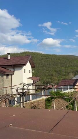 The video shows a view from a rusty rooftop looking out over residential buildings, an unplanted garden, and a heavily forested hill under a blue sky with puffy white clouds. The houses have brown roofs and white exteriors, with one house featuring brick accents. The garden area appears to be prepped for planting, with rows of tilled soil and support structures for plants. The hill is covered in dense, green trees, creating a lush backdrop. The sky is bright with scattered clouds, adding depth to the scene. The camera pans slightly, showing more of the landscape.