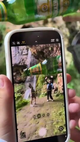 A hand holds a smartphone in landscape orientation, displaying the phone's camera interface. The camera is pointed at a scene featuring two people standing near a waterfall. A green glass bottle is positioned in the foreground, partially obscuring the view of the people and the waterfall. The bottle is held at an angle, with its neck pointing towards the right side of the frame. The camera interface shows various shooting modes, including options for photo and video. The background consists of rocks and lush green foliage. In the upper portion of the video, out of focus, are more green glass bottles. The person holding the phone is pressing the shutter button.