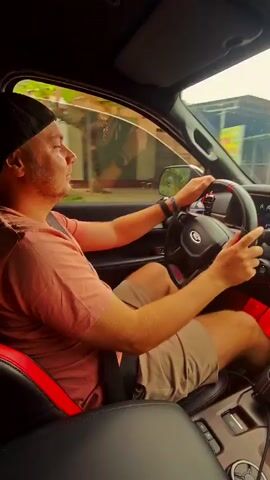 A man drives a vehicle, probably a car, on a rural road. The man is light-skinned with short hair and a beard. He wears a black baseball cap, a peach-colored T-shirt, and tan shorts.  He is strapped in with a black seatbelt. He holds the steering wheel with both hands, focusing on the road.  The car’s interior features red and black seats, and the dashboard and steering wheel show the Ford logo.  Outside, the environment consists of dense green vegetation on both sides of the road. The road appears to be winding, as the view through the windshield changes.