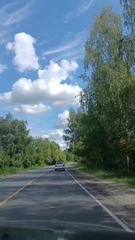 The video shows a view from inside a car driving on a two-lane road surrounded by trees. A white car is visible ahead on the road. The sky is blue with white, puffy clouds and some wispy clouds. There are power lines visible in the sky. The road appears to be asphalt with a yellow line marking the center and a white line marking the right edge. The trees on either side of the road are lush and green, indicating a summertime setting. The camera is positioned at a low angle, likely from the dashboard. The sound consists of the engine noise of the car and possibly wind. The speaker in the audio says, "I'm just a girl compared to her."