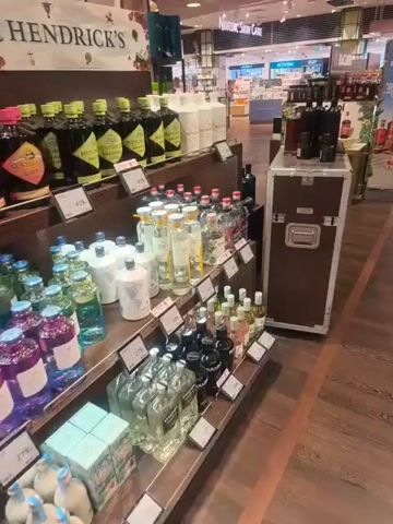 The video shows a display of various bottles of liquor, primarily gin, in what appears to be a store or duty-free shop. The bottles are arranged on shelves, with some bottles featuring a prominent Hendrick's Gin branding. The shelves are made of wood and metal, and price tags are visible on some of the shelves. In the background, other parts of the store are visible, including more shelves of products and some people walking around. There is a promotional sign for Hendrick's Gin that reads "SAVE 10% TODAY". The overall environment is well-lit and organized. The camera pans across the display, showing different brands and colors of liquor bottles.
