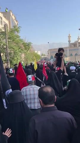 A group of people are walking down a street. Many of them are wearing black clothing, and some are wearing black headscarves. Several people are holding flags, including black flags with white images and red flags. One person in the foreground is holding a black flag with a white image of a face. Another person in the foreground is wearing a plaid shirt. In the background, there are buildings and a mosque with a minaret. The sky is blue and clear, and there are mountains in the distance. The atmosphere appears to be somber and reflective.