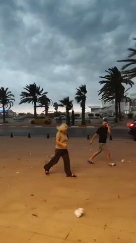 A cloudy sky looms over a beachside scene with palm trees lining a street. Two people are walking across a sandy area in the foreground. A woman with blonde hair walks from left to right, wearing a tan shirt and dark pants. A man, partially obscured, walks behind her and to the right, wearing a dark t-shirt and shorts, and is talking on a cell phone. In the background, cars are visible on the street, including a yellow taxi. Buildings with white exteriors and some vegetation are also present. The ground is covered in sand, with some debris scattered around. The lighting suggests it is either dawn or dusk.