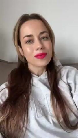 A woman with long brown and blonde hair, wearing a gray hoodie and bright pink lipstick, is seen from a close-up angle. She initially makes a kissing face, then smiles at the camera. The background appears to be a neutral wall.