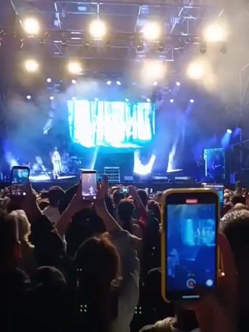 A crowd of people is recording a concert performance with their mobile phones. The crowd is mostly comprised of young adults, and they appear to be enjoying the show. The performer, a blonde woman wearing a blue dress, stands on stage in front of a large screen displaying the word "Melody" in a stylized font against a forest backdrop. The stage is lit with bright blue and white lights. A drummer is visible behind the singer. The overall atmosphere is energetic and festive.