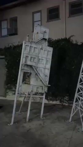 Here's a description of the video:
The video shows outdoor props on a soundstage. There is a prop of a building on stilts, a tower-like prop with a red light on top, another building prop with a water tower, and a prop of an old film camera on a tripod. There is an apartment building in the background with a wall covered in vines. It is dark out.