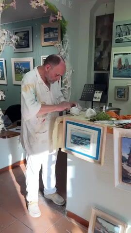 A man in a paint-splattered white smock and white pants works on a sculpture. He is holding a small tool and manipulating a white material, possibly clay or plaster, on a table. Several framed paintings are visible on the wall behind him and on the table. One painting in a wooden frame with a blue mat is in the foreground, propped up on the table. To the right of the sculpture, an artichoke and a pot of water are on the table. The environment appears to be an art studio.