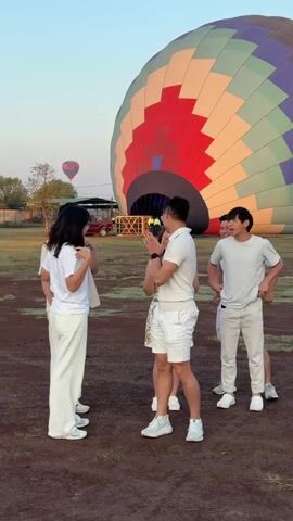 A group of six young adults stand on a patch of dry, brown dirt in front of a massive hot air balloon. The hot air balloon is patterned with large stripes in colors of red, white, purple, green, and yellow. The group is dressed in light-colored clothing, including white shirts, white pants, and white shorts. One person is wearing a floral dress. They are interacting with each other, chatting, and gesturing. There is another hot air balloon visible in the distance. A red car is parked behind a metal structure in the background. The sky is clear and pale blue. The setting appears to be a field or open area.