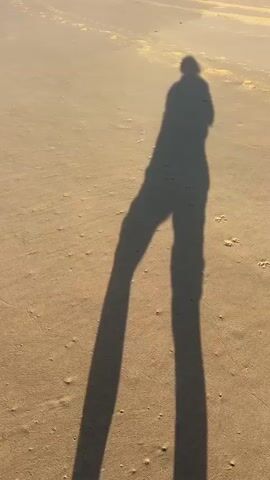 The video shows a shadow of a person on a sandy beach. The shadow is long and stretches out across the sand, indicating that the sun is low in the sky, likely at sunrise or sunset. The person appears to be standing and moving slightly, as the shadow shifts. The sand has ripples and textures visible, suggesting it's a beach environment. The lighting is warm and golden, consistent with the time of day.