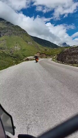 The video shows a point-of-view shot from a motorcycle or scooter traveling on a paved mountain road. In the distance, another motorcycle or scooter with a rider is visible traveling in the same direction. The road winds through a mountainous landscape with green, grassy slopes and rocky outcrops. The sky is blue with scattered white clouds. The rider's hands and a portion of the vehicle's handlebars are visible in the foreground.