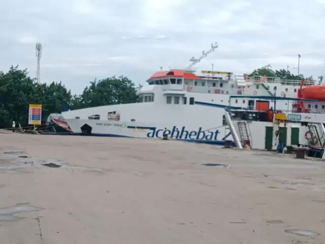 The video shows two ships docked at a port. The first ship, painted white with blue lettering, is labeled "aceh hebat" and "KMP ACEH HEBAT 3". A sign with colorful images is next to the ship. The second ship, also white, is labeled "INDONESIAN NAVIGATION SERVICE" and bears the name "AT-417 ANTARES".  Ropes connect this ship to the dock. In the background, across the water, are several large industrial buildings, including two white domed structures and a complex of towers and warehouses. The sky is overcast.