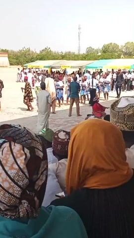 The video shows an outdoor gathering with many people. In the foreground, the backs of several individuals are visible, some wearing head coverings. In the middle ground, a group of people, predominantly women in blue and white patterned dresses, are dancing. Men in various attire, including traditional robes and casual clothing, are also present, some observing the dancers and others interacting. In the background, more people are gathered, and tents are set up. A cell tower is visible in the distance against a clear sky. The ground appears to be paved. The atmosphere seems festive.