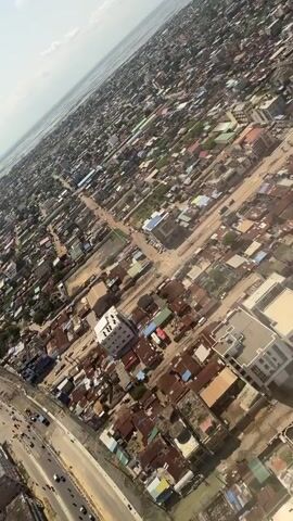 The video shows an aerial view of a densely populated urban area with numerous buildings, roads, and vehicles. The buildings appear to be of varying sizes and architectural styles, many with tin roofs. A large body of water is visible in the distance, on the horizon. Traffic flows along a wide road in the foreground. The sky is bright and clear, indicating a sunny day. The camera's perspective is angled downward, giving a comprehensive overview of the cityscape.