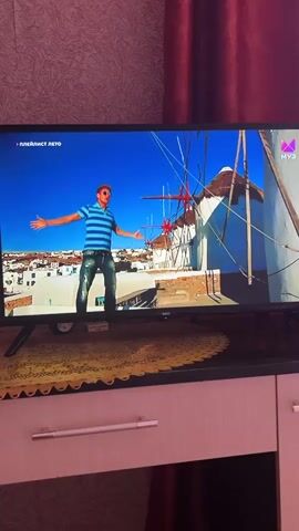 The video begins with a wide shot of a man standing with his arms outstretched in front of a white building with blue accents and a windmill in the background. He is wearing a striped blue and white shirt and blue jeans. The scene then transitions to a close-up of the man's face, with him wearing bright green sunglasses. The video then cuts to a man standing on the back of a small red truck, with his arms raised. In the background, there are white buildings and red flowers. The video appears to be a music video or a scene from a television show.