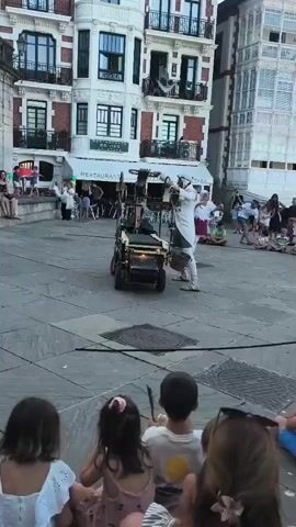 A street performer in a white robe and red sash stands in a town square, holding a long stick. He is positioned next to a small, multi-wheeled cart with a metallic frame and a lit lantern on top. The performer appears to be interacting with the cart, possibly demonstrating a skill or controlling it. A crowd of spectators, including many children, sits on the ground in a semi-circle facing the performer and the cart. In the background, buildings with ornate architecture and balconies line the square, and other people are milling about. The ground is paved with large stone tiles. The scene suggests an outdoor performance or event taking place in a European town square.