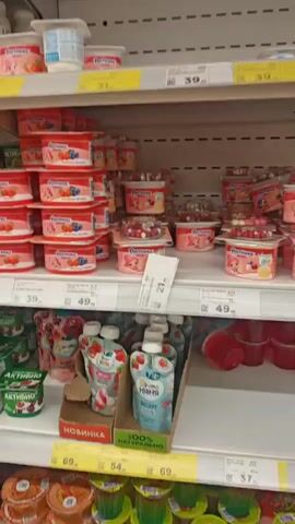 The video shows several shelves in a grocery store stocked with various dairy products, primarily yogurts and puddings. The shelves are organized by product type. In the first shot, there are multiple rows of small, individually packaged yogurts with fruit toppings. The second shot focuses on a shelf filled with a variety of yogurts and drinks, including some with cartoon characters on the packaging. The third shot displays several multipacks of Actimuno yogurt drinks. The fourth shot shows shelves of chocolate and vanilla protein puddings. The final shot pans across more shelves filled with yogurt containers and other packaged foods, and a person in a red jacket is partially visible in the foreground. The lighting appears to be typical for a supermarket, and the shelves are well-stocked.