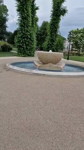 This media file is a video showcasing a park setting in Canterbury, United Kingdom. The primary subjects are a decorative fountain with water flowing and a tall monument atop a grassy hill in the background. The scene is a well-maintained park with paved pathways, lush green grass, and mature trees.

The activity taking place is a leisurely stroll through the park, with the camera capturing the environment as someone walks. The fountain is a central object, featuring a carved stone basin from which water gently cascades. Surrounding the fountain is a circular pool of water, edged with light-colored stone. Tall, slender trees with dense green foliage flank the fountain on either side. In the distance, the monument stands prominently on a rounded hill covered in grass and dotted with scattered vegetation and white wildflowers. A single bench is visible on the lawn below the monument. The architecture of buildings can be seen in the background, suggesting the park is situated near a town or city.

The time of day appears to be daytime, likely late morning or afternoon, given the soft, diffused light. The weather seems overcast, with a cloudy sky visible, contributing to a gentle and serene atmosphere. No people are clearly discernible as active participants, though silhouettes might be present in the distance near the monument. There is no visible text in the media file. The overall mood conveyed is peaceful and tranquil, highlighting the natural beauty and man-made structures of the park.