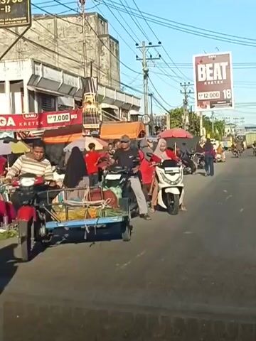 The video shows a street scene with several people and vehicles. In the foreground, a person is riding a white scooter, and another person is on a motorcycle with a trailer. There are several market stalls set up along the side of the road, with vendors selling fruits and other goods. A large billboard advertising "Beat Kress" is visible in the background. The weather appears to be sunny. The people are dressed in casual clothing, and some are wearing headscarves. The overall impression is of a busy street market.
