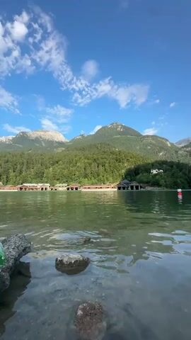 The video shows a body of water with rocks in the foreground. In the background, there is a shoreline with several buildings that appear to be boathouses, nestled amongst trees. Beyond the trees are mountains, one of which is partially snow-capped. The sky is blue with some white clouds.