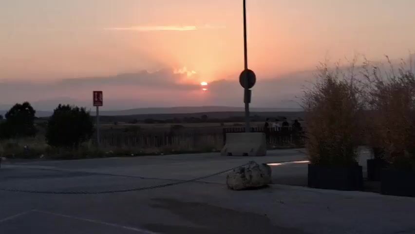 The video shows a sunset over a rural landscape. The sun is low in the sky, casting a warm orange glow. There are some clouds in the sky, which are lit up by the sun's rays. In the foreground, there is a road with a chain and a large rock on it. There are also some people standing on the side of the road, watching the sunset. A lamppost with a sign is visible. There is also a fire exit sign on the left side of the video.