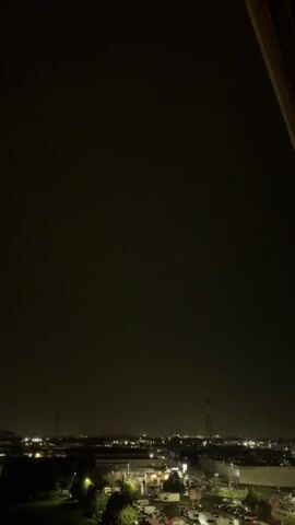The video captures a nighttime cityscape with a dark, cloudy sky. A powerful lightning strike illuminates the sky with a bright flash, briefly revealing the outlines of buildings, streetlights, and what appear to be power line towers. The lightning is a vivid, jagged line across the upper part of the frame. After the flash, the scene returns to darkness, punctuated by the distant lights of the city.