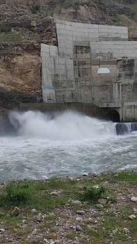 The video shows a large, imposing concrete dam constructed on a steep, rocky mountainside. The dam is a massive structure with many tiers and sections, appearing to be either under construction or recently completed, as indicated by exposed concrete and some graffiti on its surface. Water is being released from the dam in a powerful surge, creating a large plume of white spray and foam that crashes into the turbulent river below. The river is wide and fast-flowing, with greenish-gray water churned up by the force of the release. The surrounding landscape is rugged and natural, with the brown, rocky slopes of the mountain forming a stark contrast to the man-made structure of the dam. A small patch of green grass and pebbles lines the bank of the river in the foreground. The lighting suggests it is daytime, with overcast or diffused light, as there are no strong shadows. The overall impression is one of immense power and the force of nature interacting with human engineering. The visible text on the dam is graffiti, some of which is illegible.