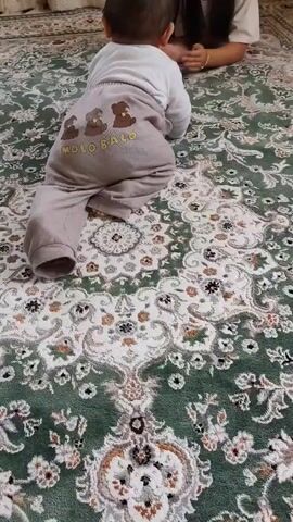 A baby wearing a white shirt and light brown pants with bear graphics on the back is crawling on a patterned green rug. The baby's back is to the camera. Another person, a young woman, is sitting on the rug in the background, facing the baby. She has dark hair and is wearing a white shirt. Her hands are clasped together. The woman appears to be watching and encouraging the baby. The rug has an ornate, traditional pattern with floral motifs and a central medallion.