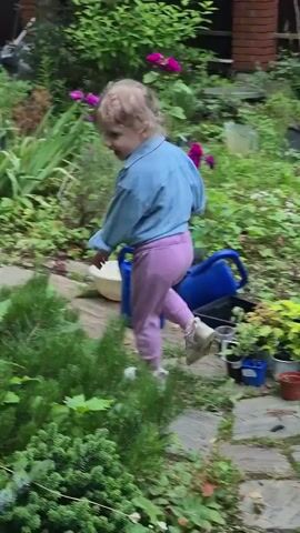 A young child, wearing a blue denim jacket and pink pants, walks along a paved path in a lush garden. The child has blonde, curly hair and appears to be smiling. In the background, there are various green plants and flowers, including some with bright pink blossoms. A blue watering can and a black planter are visible to the right of the path. Further back, a white vehicle is parked near a building with red brick accents. The scene suggests a daytime setting, possibly in the afternoon, with fair weather suitable for outdoor activities. The environment indicates a residential area with a well-maintained garden.