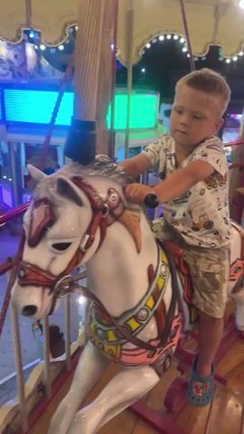 A young boy rides a white carousel horse with red and gold accents. He wears a white t-shirt with a graphic print and camouflage shorts. The carousel is illuminated with colorful lights, including blue and green lights in the background. The boy is holding onto the reins and appears to be looking slightly off-camera. The carousel horse has a detailed mane and bridle.