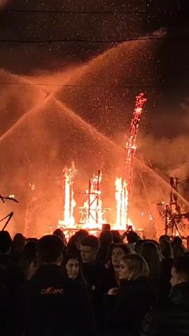 A large wooden structure burns intensely amidst a crowd of people at night. Two streams of water are aimed at the fire, sending droplets into the air. The fire illuminates the faces in the crowd, which consists of men and women of varying ages, watching the spectacle. Some are looking directly at the fire, others are engaged in conversation. The light from the fire casts long shadows, and the silhouettes of the people stand out against the bright flames. The background is dark, with power lines visible above.
