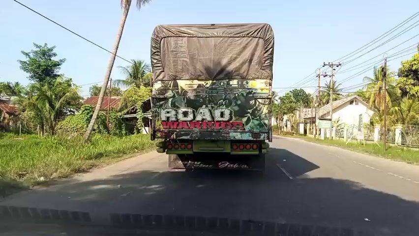 A rear view of a truck driving on a paved road. The truck has a black tarp covering its cargo area. The back of the truck is decorated with a colorful graphic and the words "ROAD WARRIORS" in large, bold letters. The truck has multiple red and yellow lights on its rear. The road is lined with green grass and vegetation on the left, and houses and palm trees on the right. The sky is clear and blue. The video appears to be recorded from a moving vehicle behind the truck.
