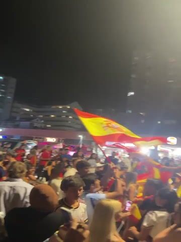 The video shows a large crowd of people at night, waving Spanish flags and cheering. The crowd is densely packed, and many people are holding up their phones to record the scene. Buildings are visible in the background, and there are streetlights illuminating the area. Some individuals in the crowd appear to be on the shoulders of others, and some are wearing Spanish flags as capes. The atmosphere seems celebratory, likely related to a sporting event or national holiday.