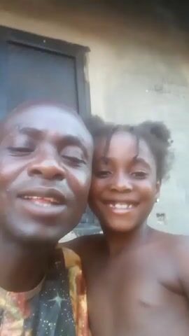 A man with short hair and a dark complexion, wearing a colorful shirt, is holding a camera to take a selfie with a young girl with braided hair. The girl stands beside him, bare-chested. Both are smiling. They are positioned in front of a building with a dark door and a light-colored wall that has some scribbling on it. The man expresses his love for the girl.