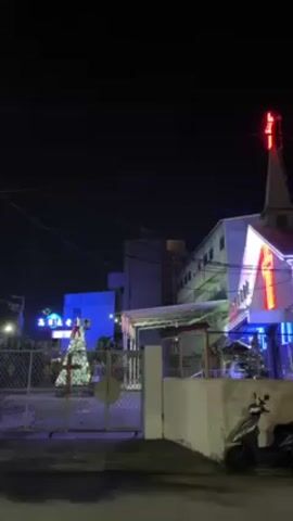 The video shows a nighttime scene of a church. The church building is brightly lit with blue and red neon lights, including a red cross at the top of its steeple. In front of the church, there is a chain-link fence, and behind the fence, a Christmas tree is decorated with white lights. To the right of the fence is a small white wall and a parked scooter. The sky is dark, and utility lines are visible stretching across the frame. In the background, additional buildings can be seen, lit with blue lights. The scene is still and quiet.