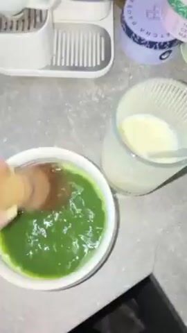A person is making a matcha latte. A small, white bowl containing a dark green liquid is on a grey counter. A person’s hand holding a wooden whisk is seen whisking the liquid in the bowl. Next to the bowl is a clear glass holding a pale yellow liquid with a spoon in it. In the background are two round, purple and green cans. A white coffee maker is also in the background to the left of the cans.