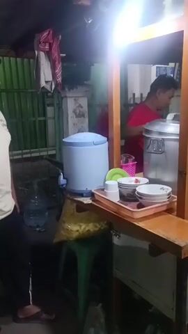 The video shows a food stall at night. A man stands on the left, facing away from the camera, wearing a light-colored shirt and dark pants. The stall is illuminated by a bright light hanging overhead. On the counter, there is a blue water dispenser, a tray with bowls and plates, and two large metal pots. A person in a red shirt is visible behind the counter, attending to the food. The stall has a wooden frame and a wooden counter. In the background, there is a green fence and a large water bottle. The overall environment appears to be a street or an outdoor food court.