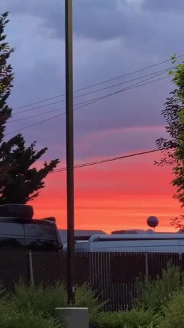 This vertical video captures a vibrant sunset over a suburban setting in Milton, USA. The sky is a dramatic display of color, transitioning from a deep purple at the top to fiery orange and red near the horizon. Wispy clouds are scattered across the sky, catching the light and adding depth to the scene.

In the foreground, a chain-link fence encloses a yard with lush green bushes. Behind the fence, several objects are visible. On the left, a dark-colored vehicle, possibly an SUV, has a spare tire mounted on its roof. To the right of the vehicle, the roof of a white van or recreational vehicle can be seen. Perched atop this vehicle is a single, dark-colored balloon, adding an unexpected and slightly whimsical element to the composition. A tall, dark pole stands prominently in the center of the frame, obscuring a portion of the sky and the objects below. Power lines stretch across the upper part of the image, intersecting with the colorful sky. Green trees frame the right side of the scene, their leaves illuminated by the setting sun.

The time of day is clearly sunset, indicated by the intense colors in the sky and the long shadows that would be cast. The weather appears calm, with the clouds suggesting a beautiful end to the day rather than an impending storm. The overall emotion evoked is one of awe and tranquility, with a hint of playful curiosity due to the balloon. No visible text is present in the media file.