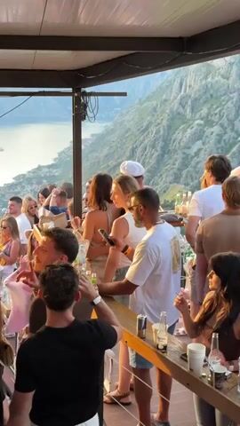 The video shows a crowded outdoor party at dusk with a scenic mountainous coastline and body of water in the background. People are standing and socializing around tables and a bar. Many are holding drinks and looking at their phones. A speaker is suspended from the ceiling. The lighting suggests a golden hour ambiance.