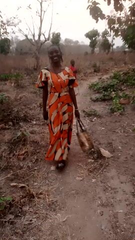 A woman with dark skin stands on a dirt path in a rural area. She is smiling and holding a broom behind her back. She is wearing a long orange dress with a leaf-like pattern. The ground is covered with dry leaves and small plants. There are trees on either side of the path. The video is vertical.