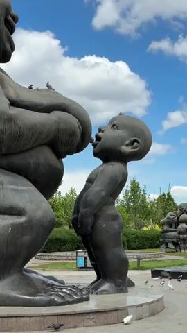 The video shows a close-up view of a sculpture depicting a mother breastfeeding a child. The sculpture is made of a dark, possibly metallic material and is set in a park. The mother figure is large and squat, with exaggerated features and a bun on top of her head. The child figure is smaller and stands facing the mother, with its mouth open and attached to the mother's breast. Several pigeons are perched on the sculpture, on the head of both mother and child. The background includes green trees, a blue sky with white clouds, and other sculptures in the distance. A trash can is located on the left side of the sculpture, near the base. The camera moves slightly, circling the sculpture to show different angles.
