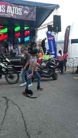 The video depicts a street scene with a woman dancing in front of a stage. The woman is wearing jeans, a patterned shirt, a hat, and boots. She is dancing with her arms outstretched. There is a band performing on stage behind her, with lights and banners visible. Several motorcycles are parked near the woman, and other people are standing around watching. The ground appears to be asphalt.